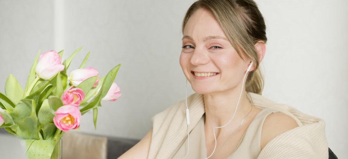 A cheerful woman wearing headphones smiles next to a vase of pink tulips indoors.