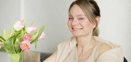 A cheerful woman wearing headphones smiles next to a vase of pink tulips indoors.