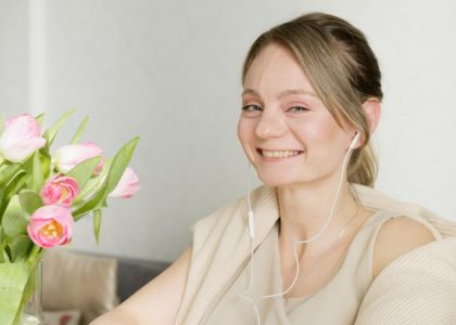 A cheerful woman wearing headphones smiles next to a vase of pink tulips indoors.